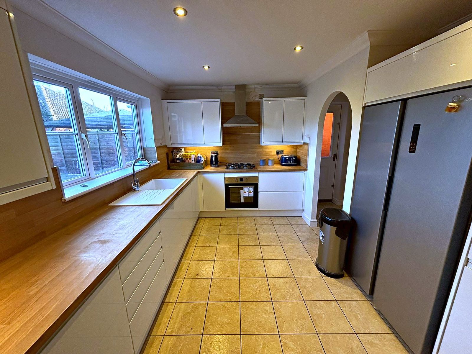White gloss kitchen with oak worktops running beneath a large window and an arched doorway