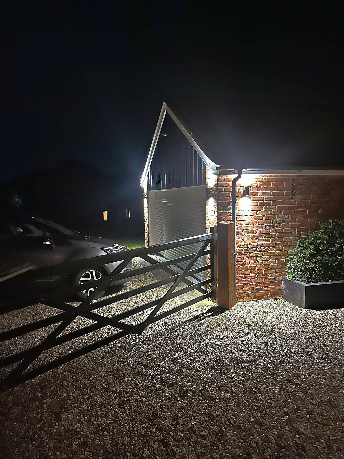 Five-bar gate, gravel drive and clad gable lit at night by exterior lighting
