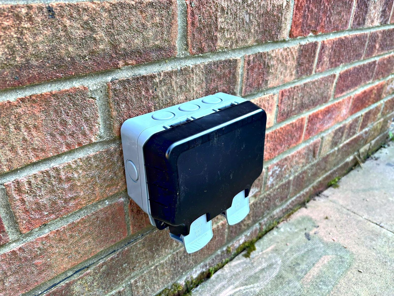 Weatherproof outdoor socket enclosure mounted on a brick wall beside a patio
