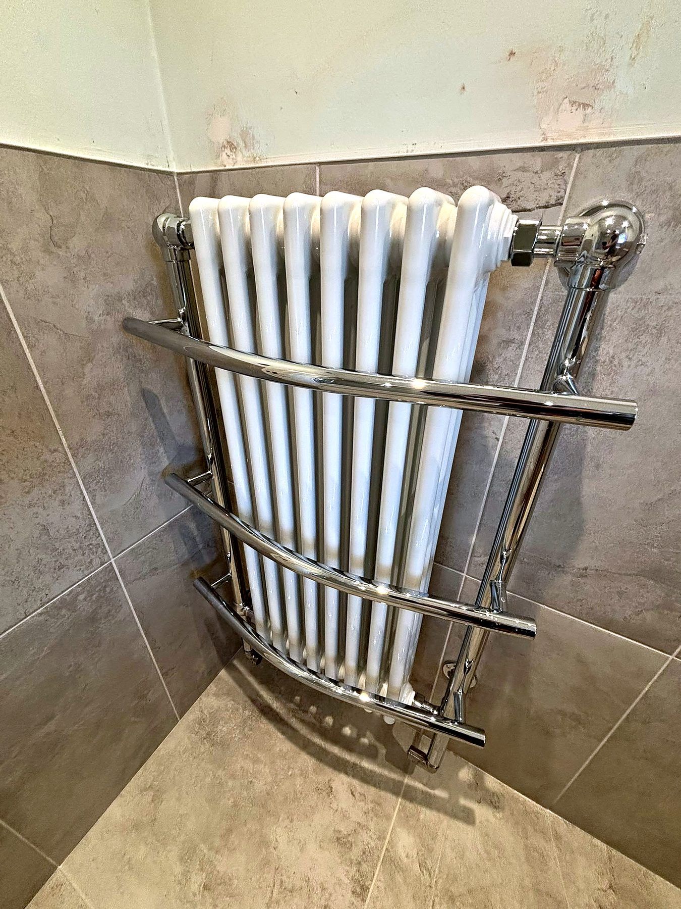 Chrome column towel radiator fitted into the corner of a grey tiled bathroom