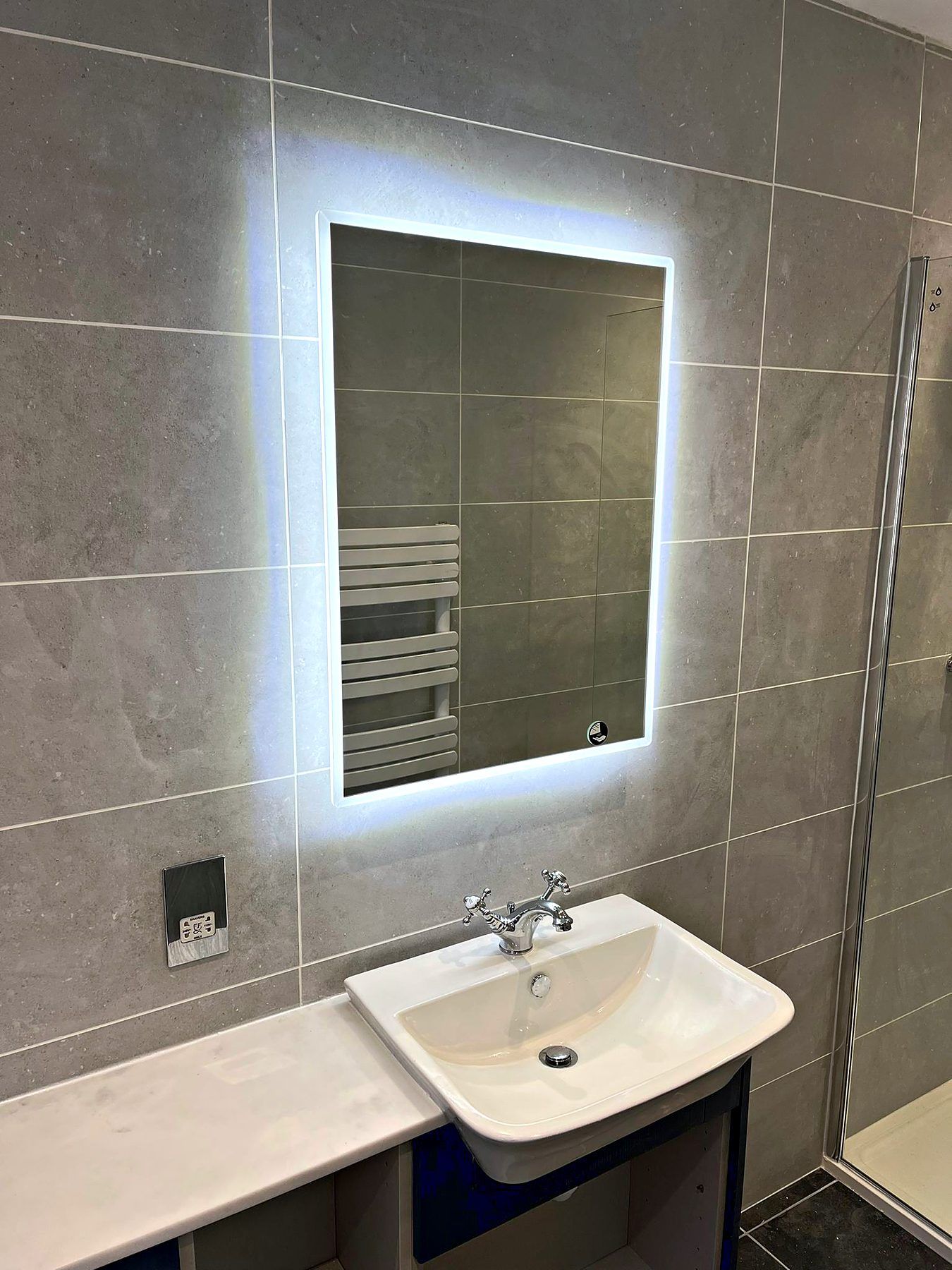 Illuminated LED mirror above a basin and vanity unit in a grey tiled bathroom